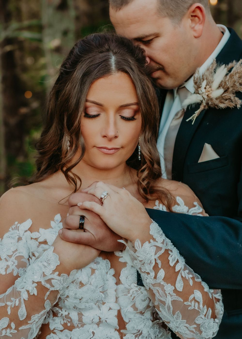 Groom embraces bride from behind as she looks down at her ring in romantic forest wedding portrait.