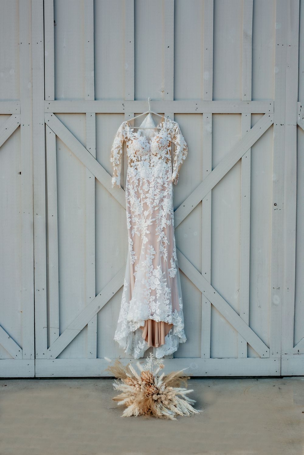 Floral lace wedding dress with flutter sleeves hanging on rustic blue barn doors with pampas grass bouquet.