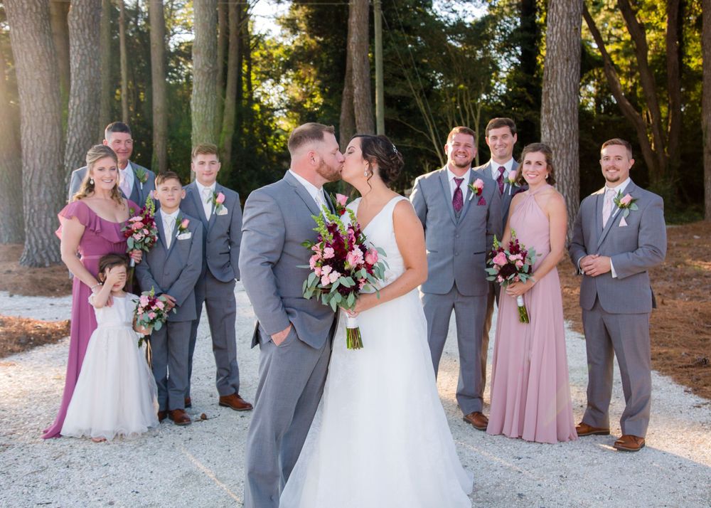 Wedding party poses together with bride and groom kissing, bridesmaids in mauve dresses and groomsmen in gray suits.