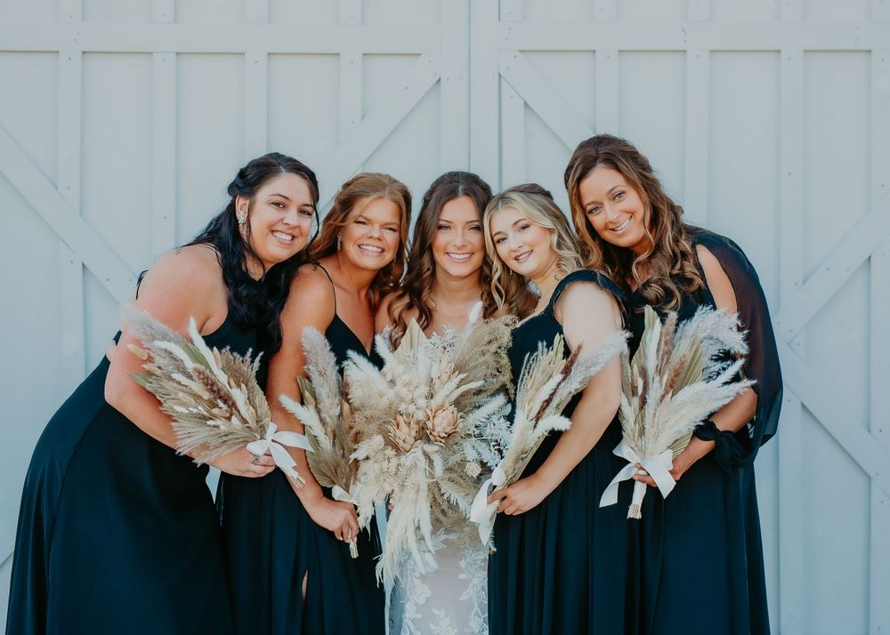 Bride with four bridesmaids in navy dresses holding pampas grass bouquets in front of white barn doors.