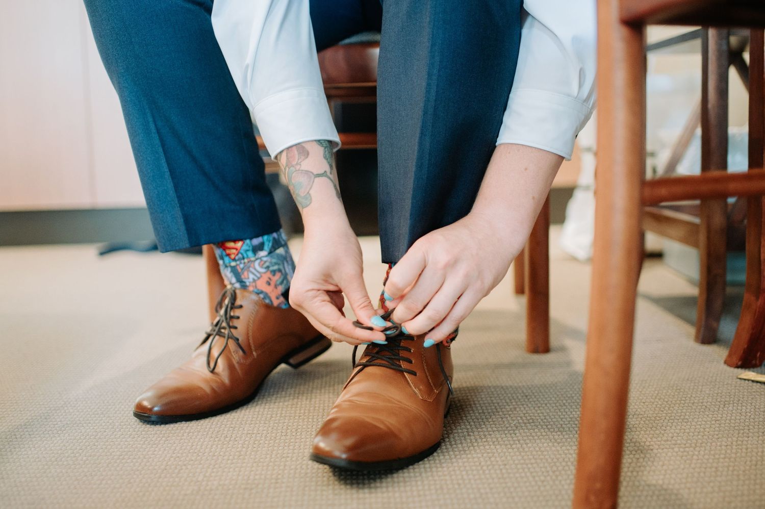 Close-up of hands tying brown leather dress shoes with visible tattoo and blue pants.