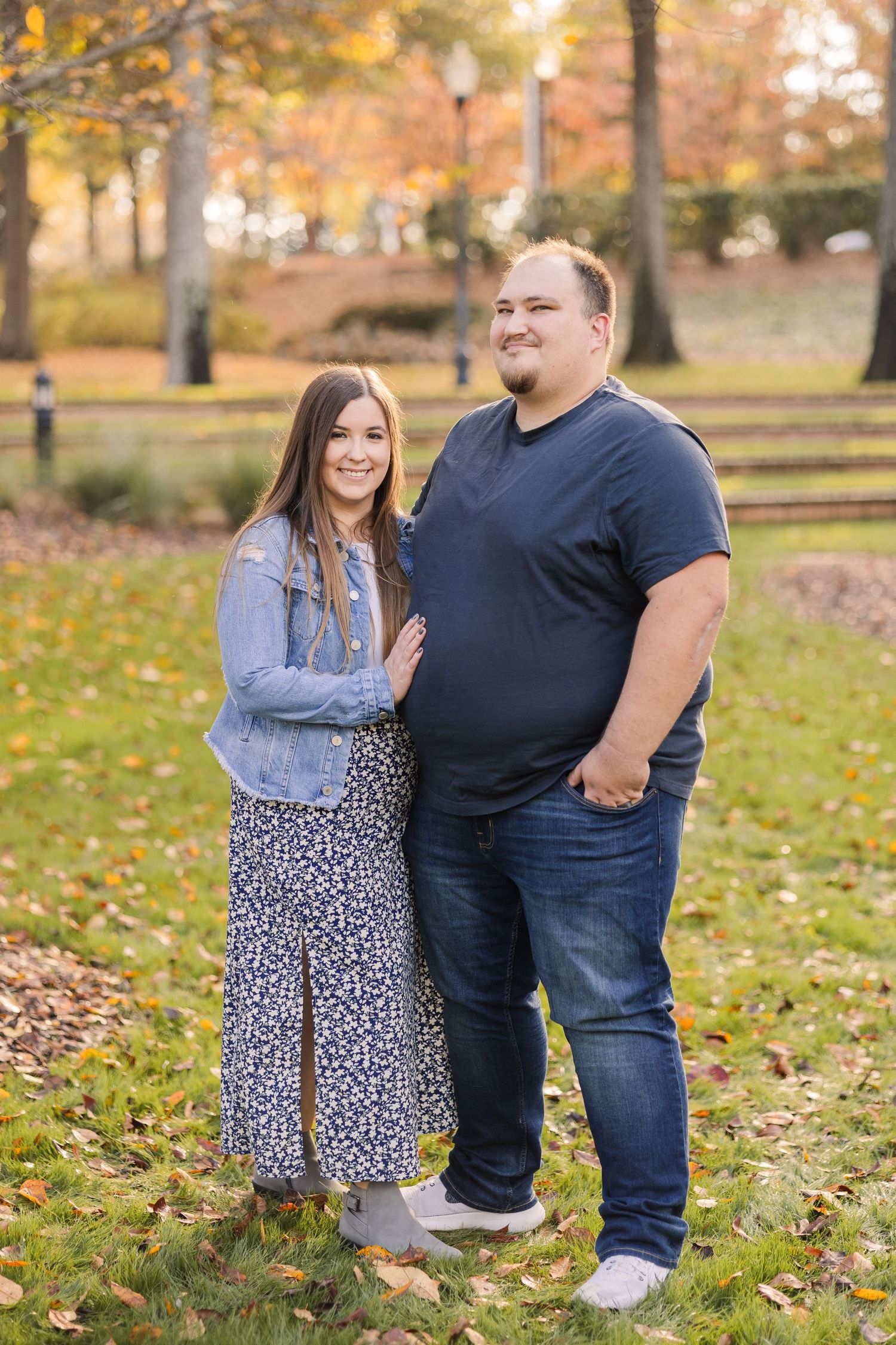A couple stands together on fallen leaves in an autumn park setting with warm lighting.