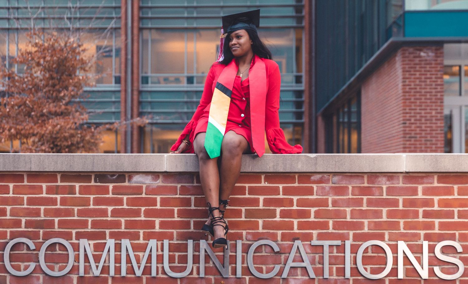 Graduate in red gown sitting on brick wall with Communications text overlay.