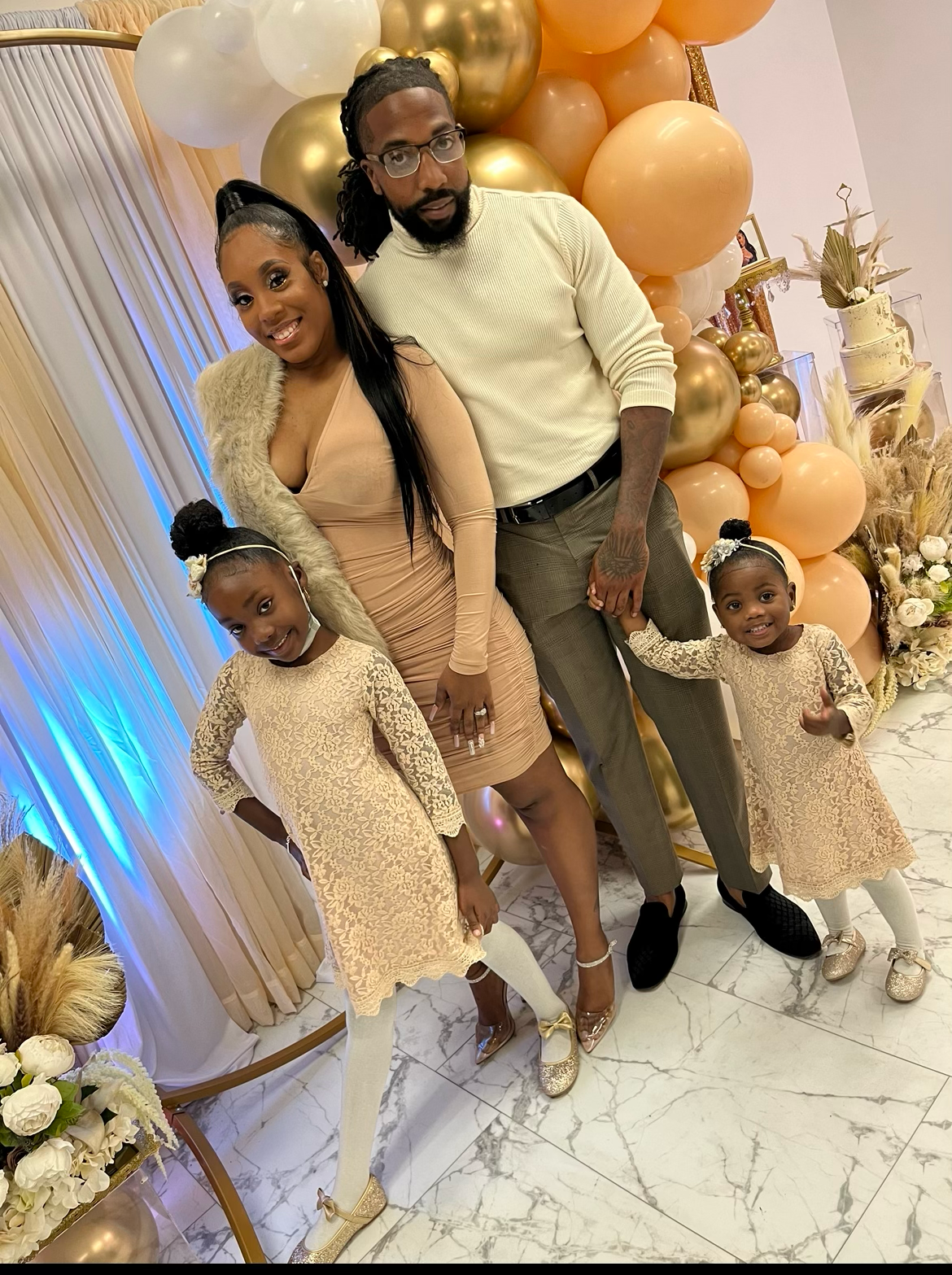 A coordinated family posing together in neutral-toned outfits against a festive gold balloon backdrop.