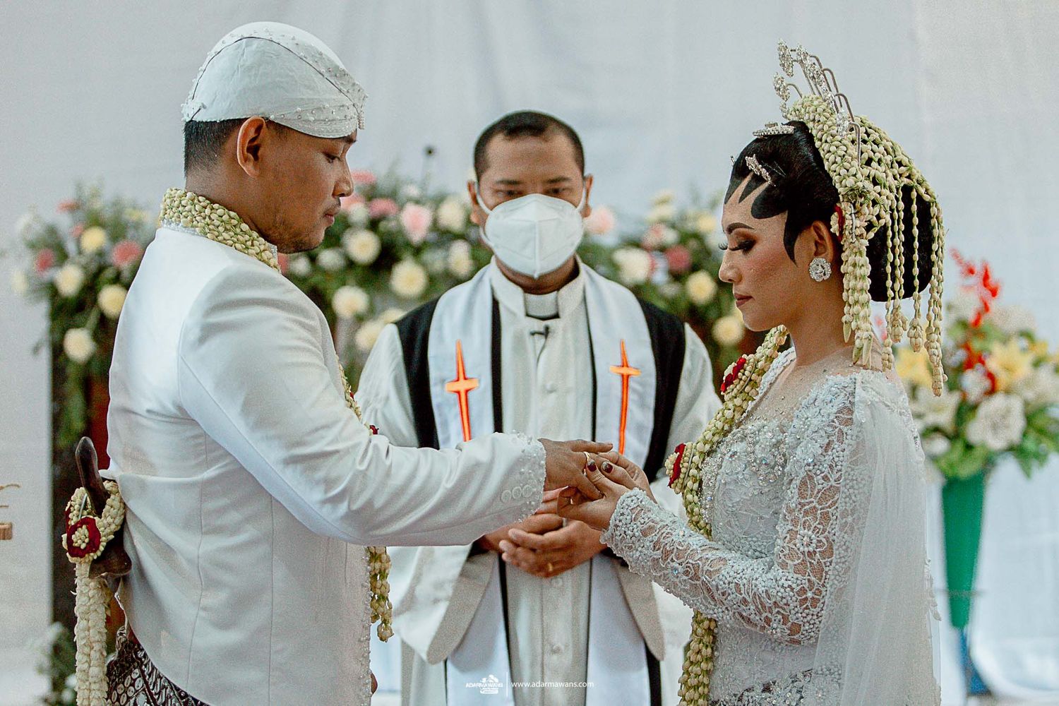 Traditional wedding ceremony with couple exchanging vows in front of officiant surrounded by floral decorations.