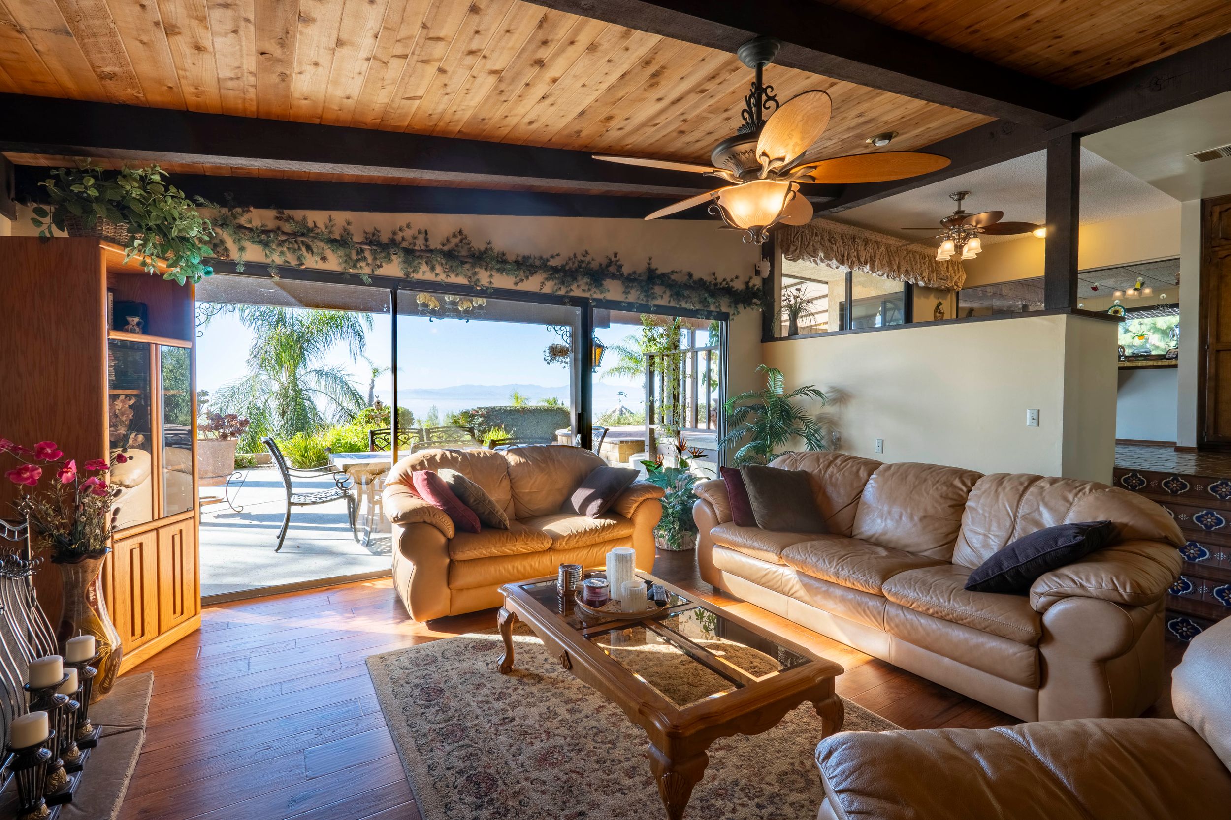 Sunlit living room with leather sofas, wood ceiling, ceiling fan, and sliding glass doors opening to a pool and ocean view.