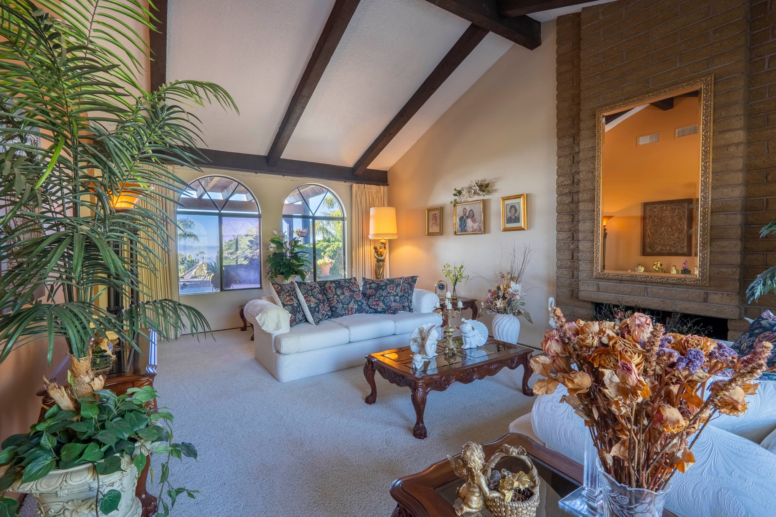 Elegant living room with vaulted beam ceiling, brick fireplace, arched windows, white sofas, and lush indoor plants.