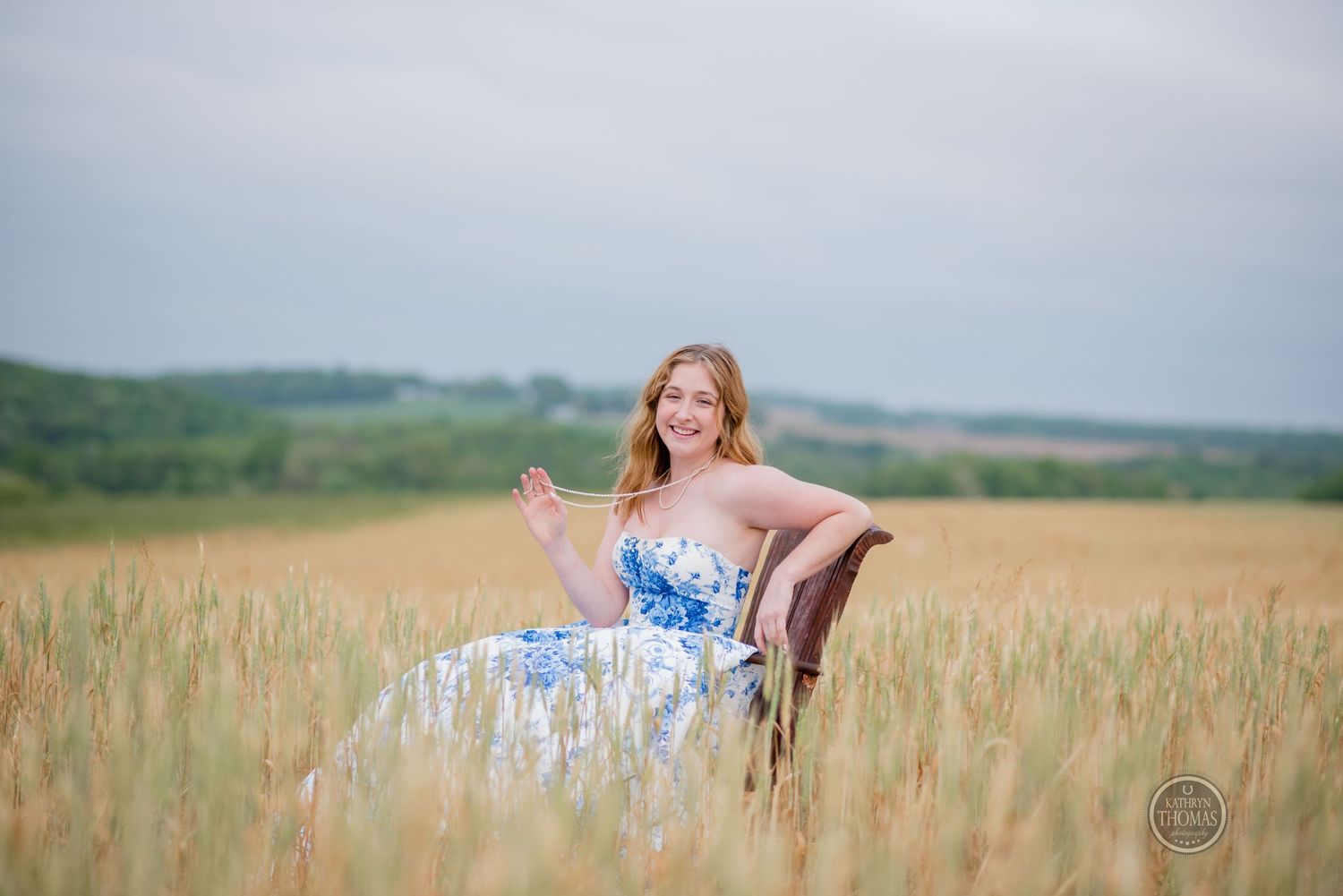 Woman wearing a floral blue gown sits in a vintage chair surrounded by tall wheat stalks.