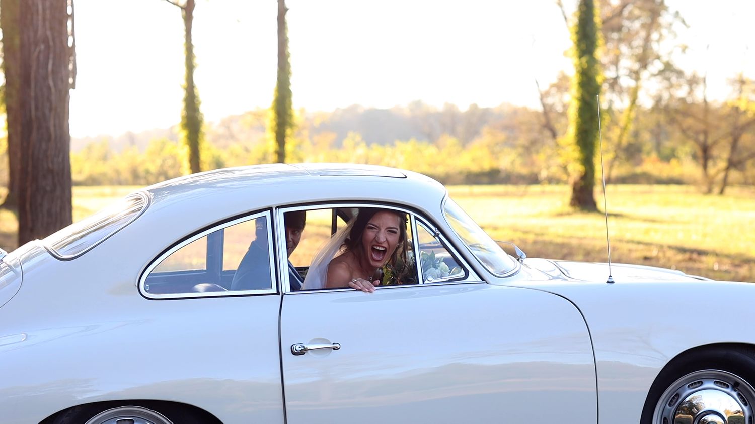 Joyful bride laughing out of window of vintage white classic car on sunny day.