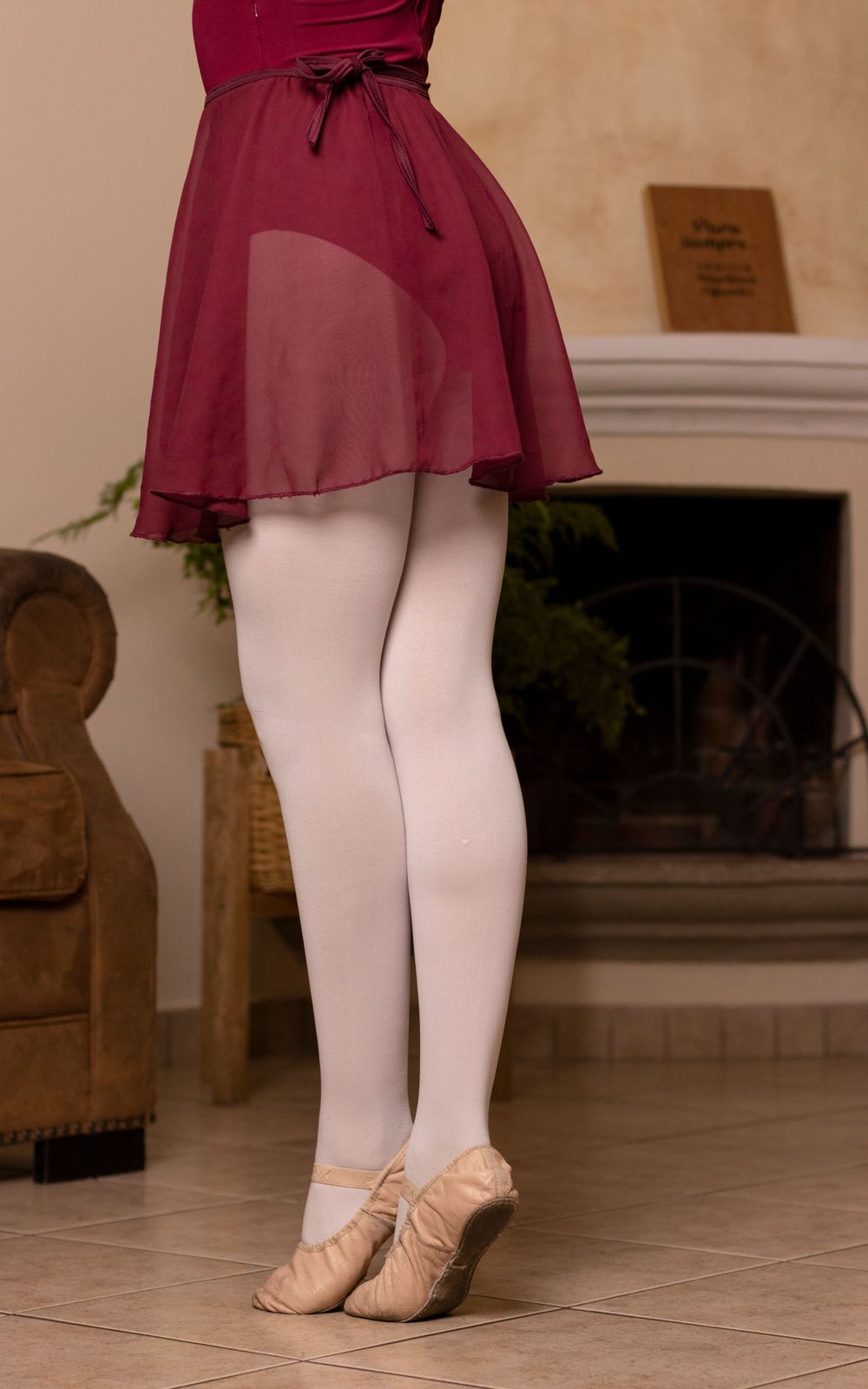 Ballet dancer in burgundy dress practicing positions with white tights on beige floor.