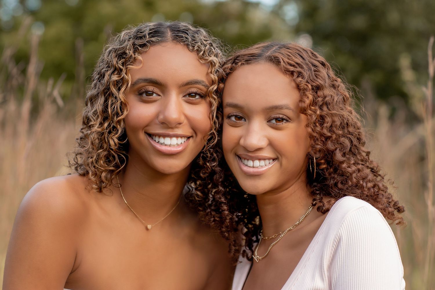 Two smiling young women with curly hair pose together outdoors in a sunny field.