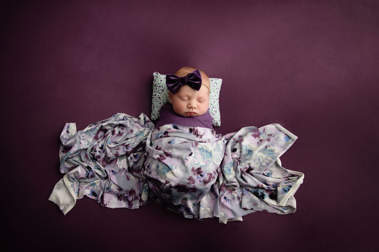 Sleeping newborn wrapped in floral swaddle on purple backdrop with black bow headband.