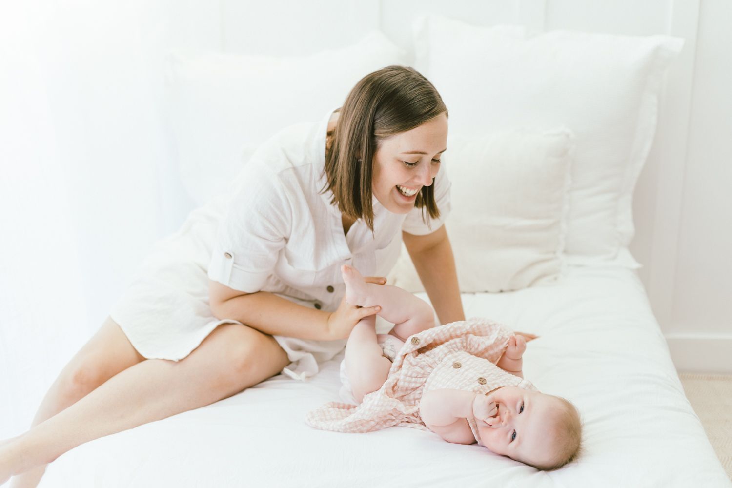 Mother plays with baby during lifestyle photography session on white bedding.