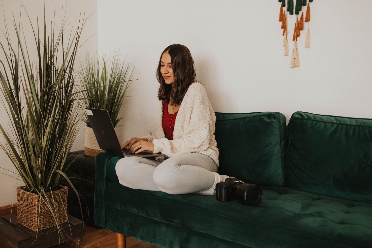 Content creator working on laptop while sitting on emerald green velvet couch with plants nearby.