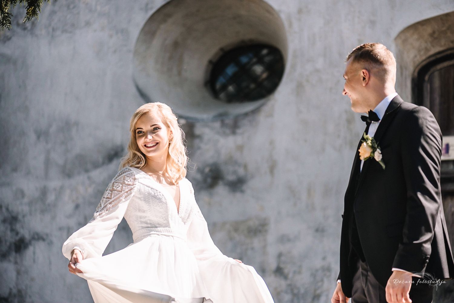 Jaunoji šypsosi apšviesta aštrios saulės, jaunikis meiliai į ją žiūri vestuvinės fotosesijos metu