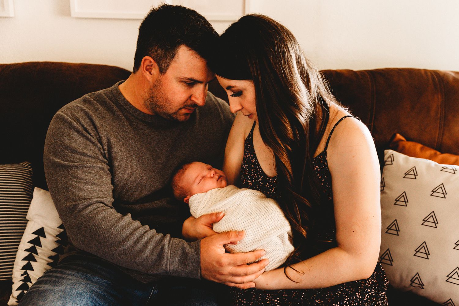 Tender newborn portrait of new parents cradling their sleeping baby on a cozy living room couch.