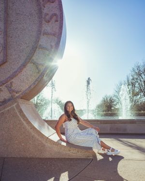 A person in a checkered dress sits on a curved stone structure near a fountain on a sunny day senior graduation portrait session