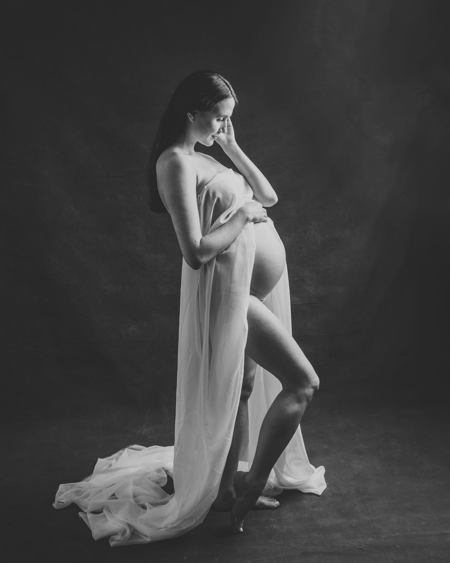 Black and white artistic maternity photo of pregnant woman in sheer white draped fabric.