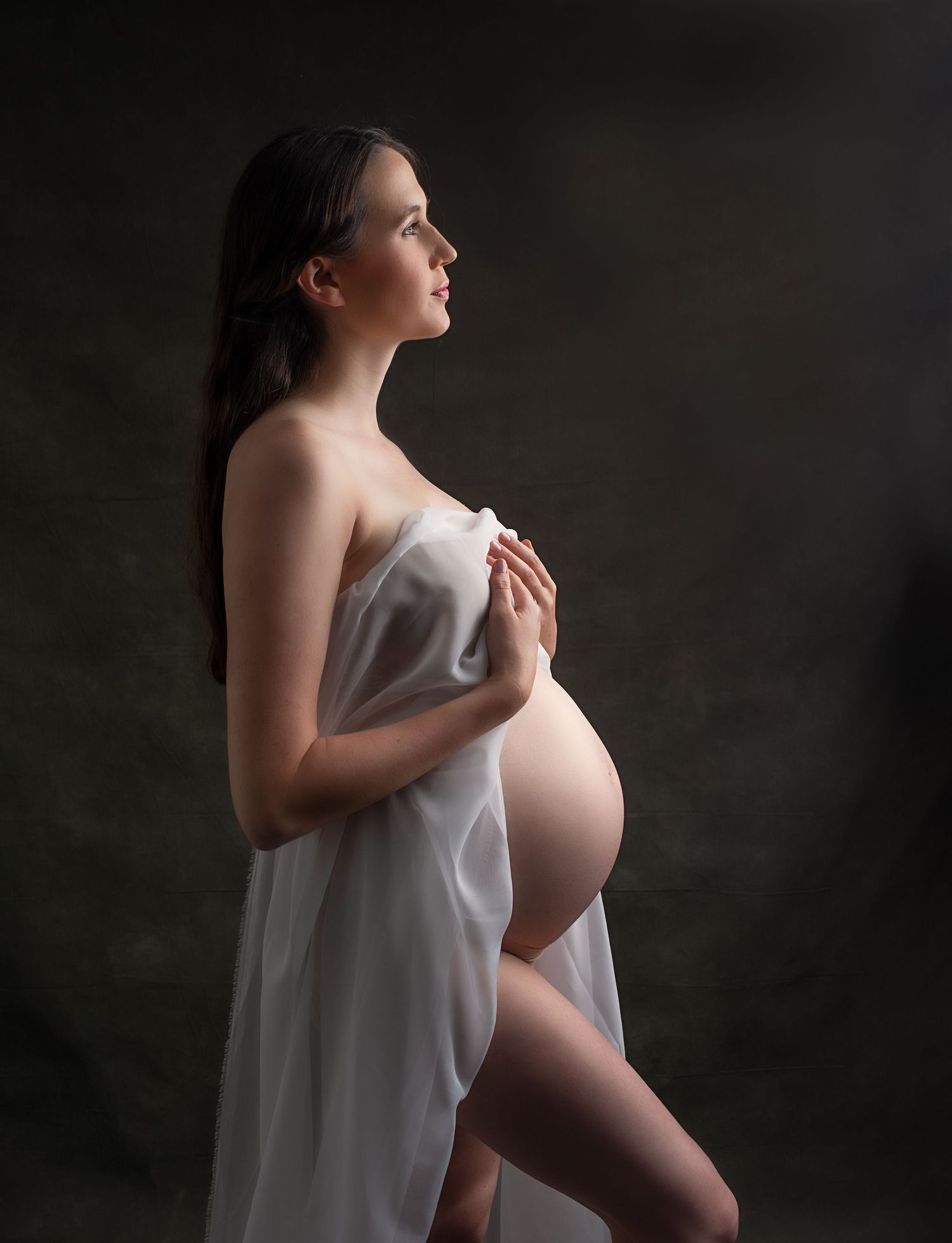 Pregnant woman draped in white fabric holding baby bump in artistic studio maternity photo.
