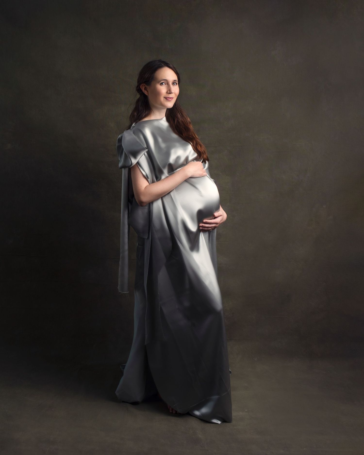 Elegant pregnant woman in full-length silver satin dress holding baby bump in studio portrait.