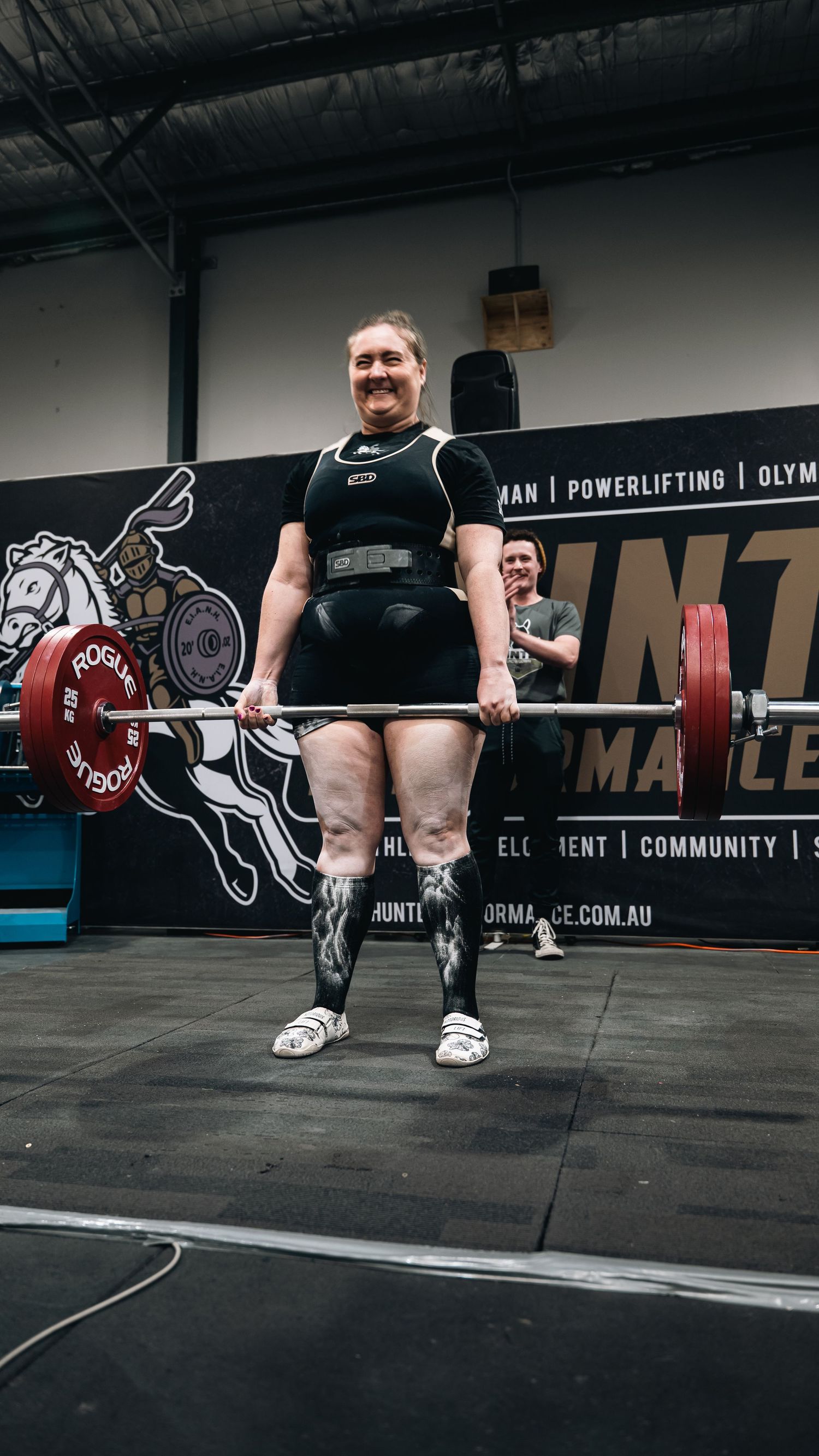 Powerlifter performing heavy deadlift at a strength competition with gym branding wall.