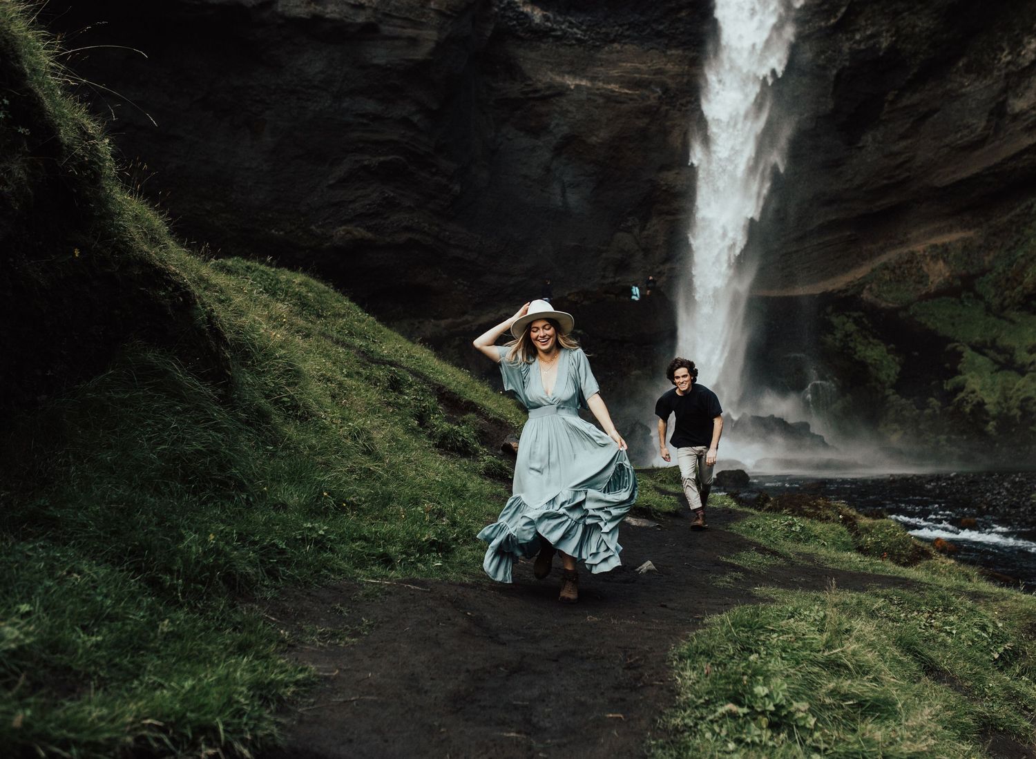 engaged couple running through green iceland landscape waterfall behind