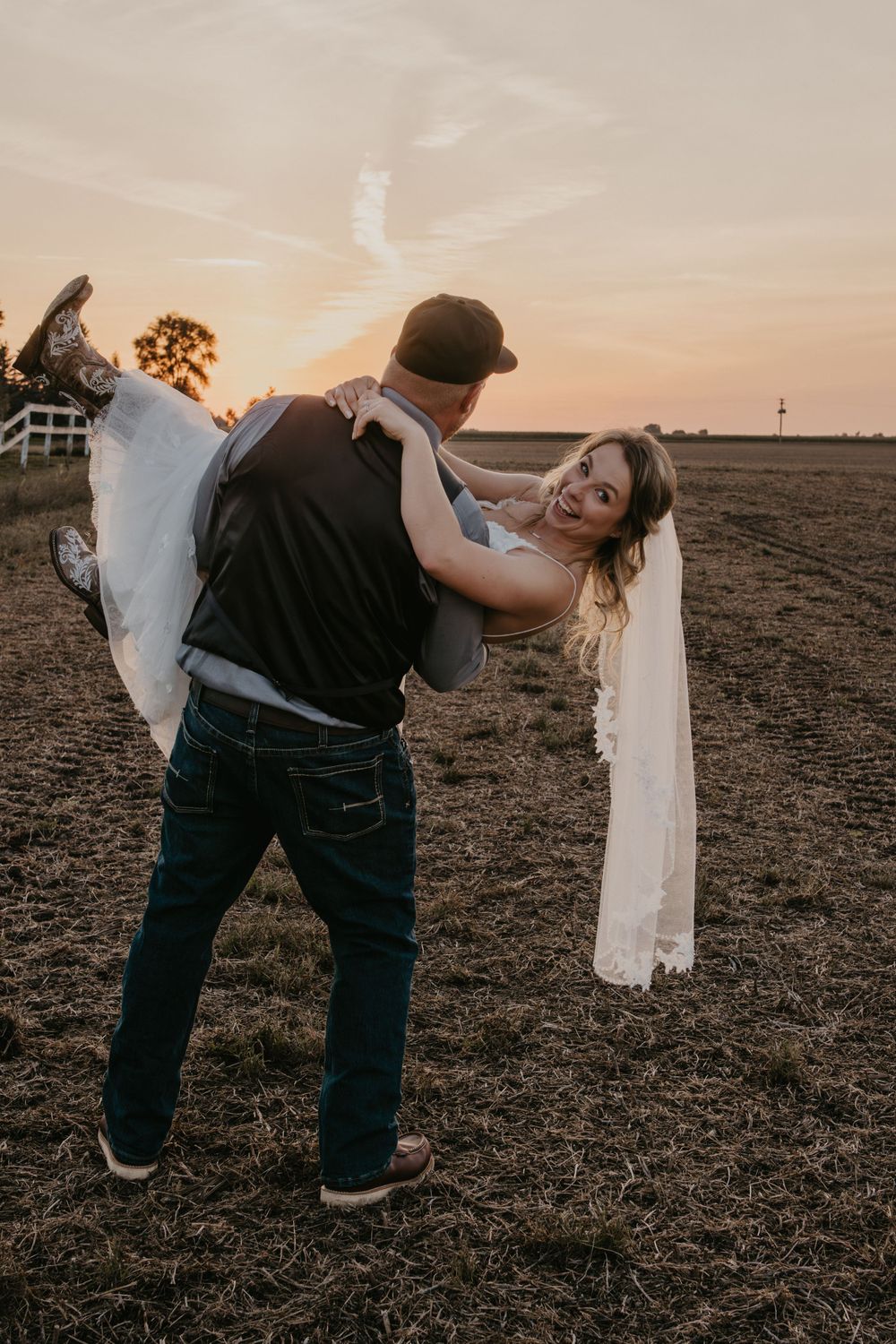 Groom lifting laughing bride in wedding dress at golden sunset over open farm field.