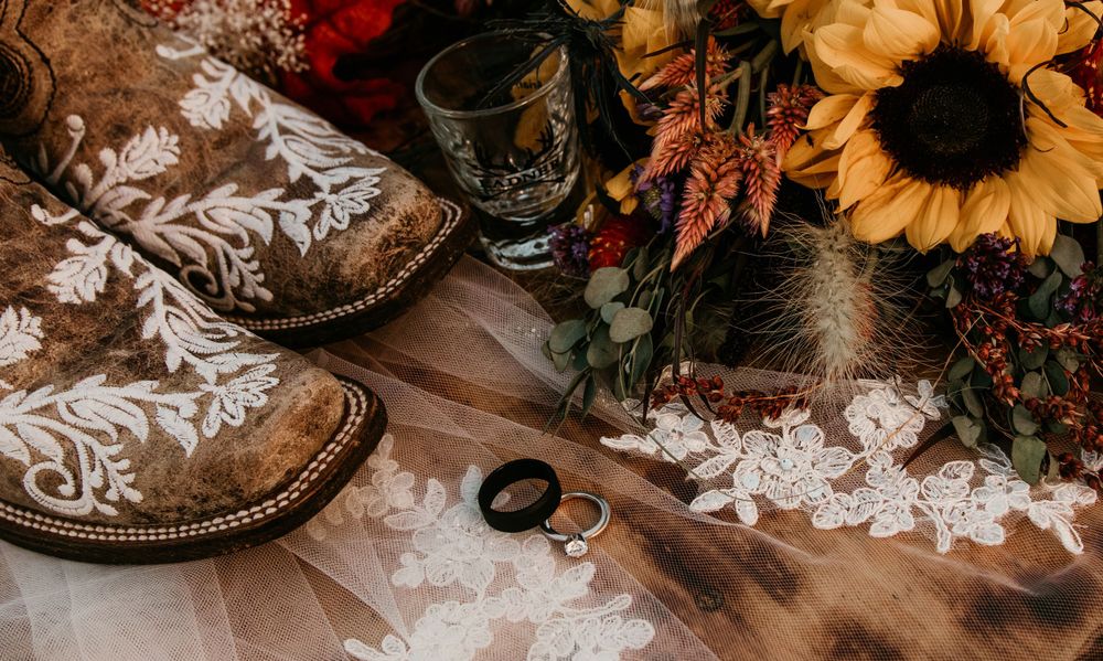 Rustic wedding flat lay featuring cowboy boots, sunflowers, fall foliage, wedding rings on lace fabric.