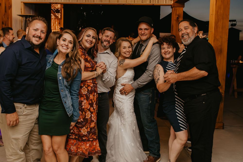 Wedding party group photo with bride in white gown smiling with family at rustic barn reception.