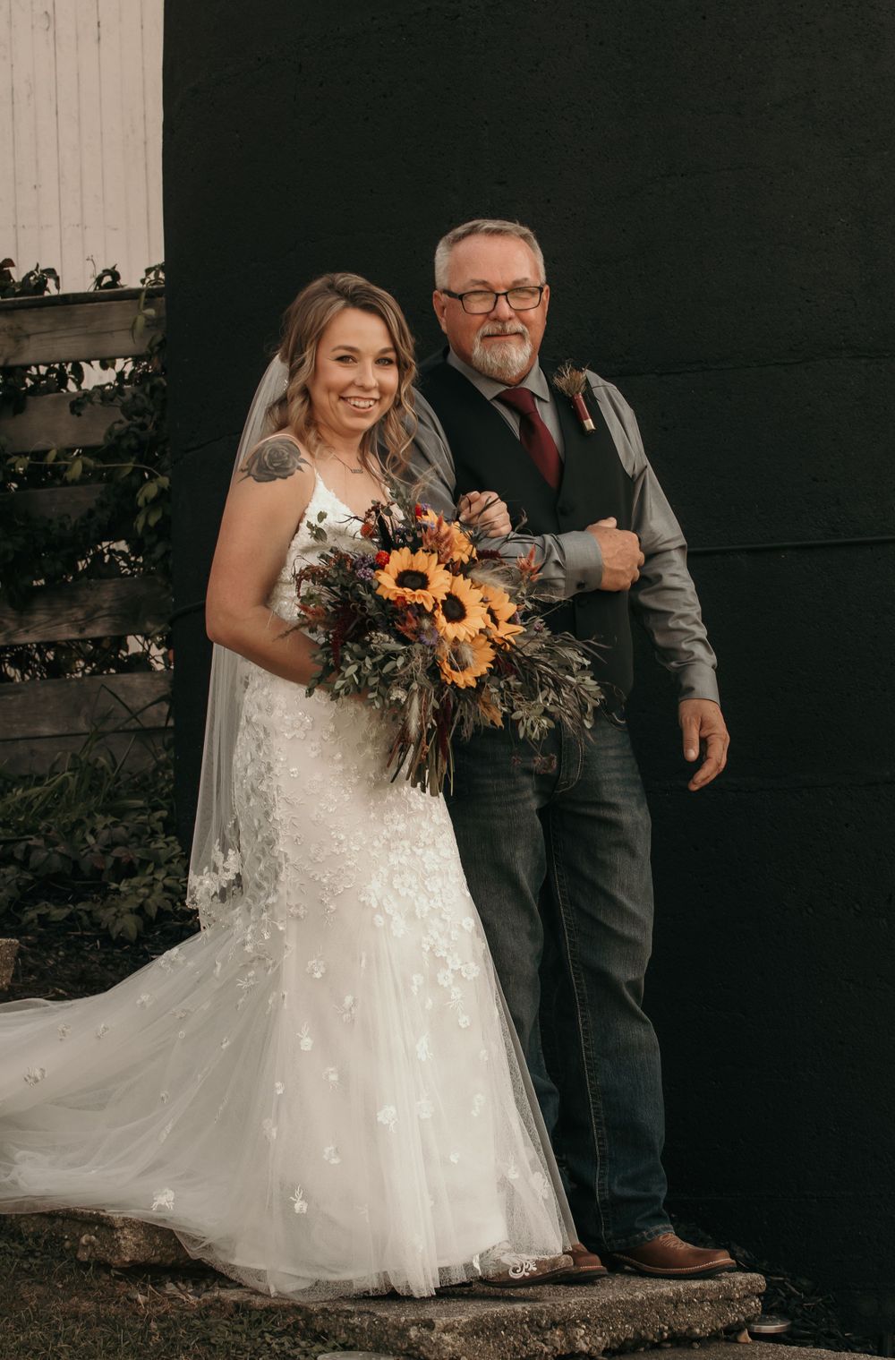 Bride in lace wedding gown holding sunflower bouquet poses with father outside black barn.