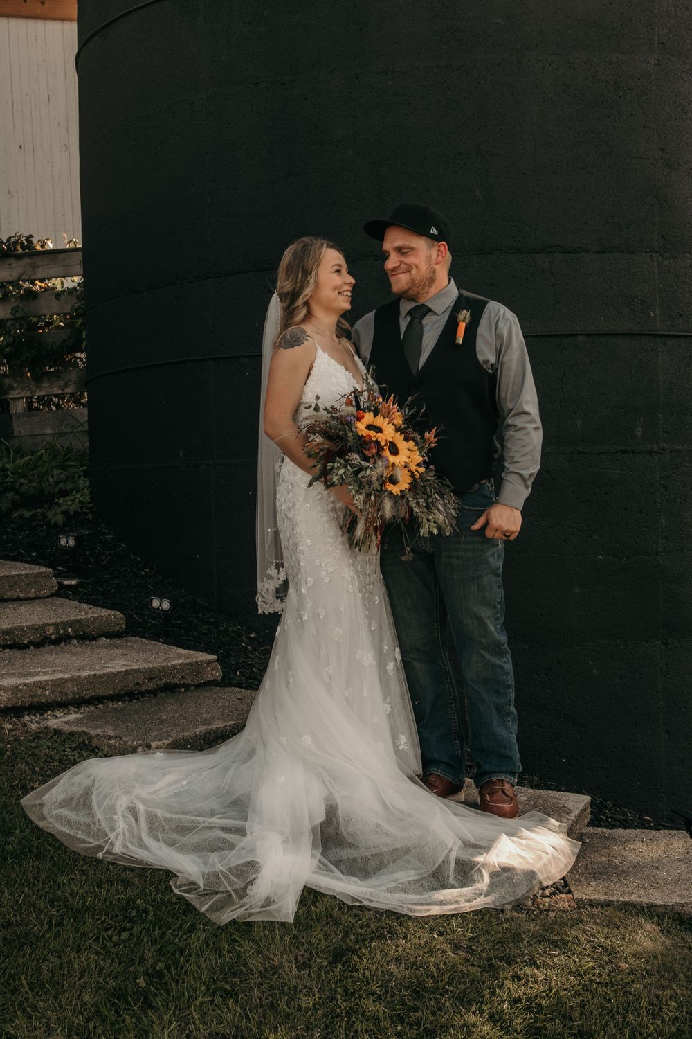 Bride and groom share a tender moment outside black barn with sunflower bouquet and veil.