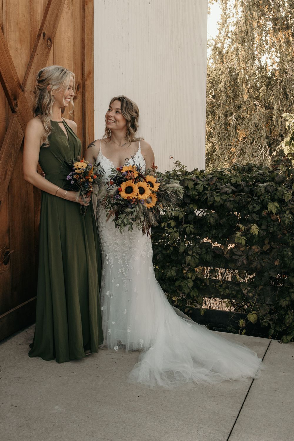 Bride in lace gown and bridesmaid in olive green dress holding sunflower bouquets by barn door.
