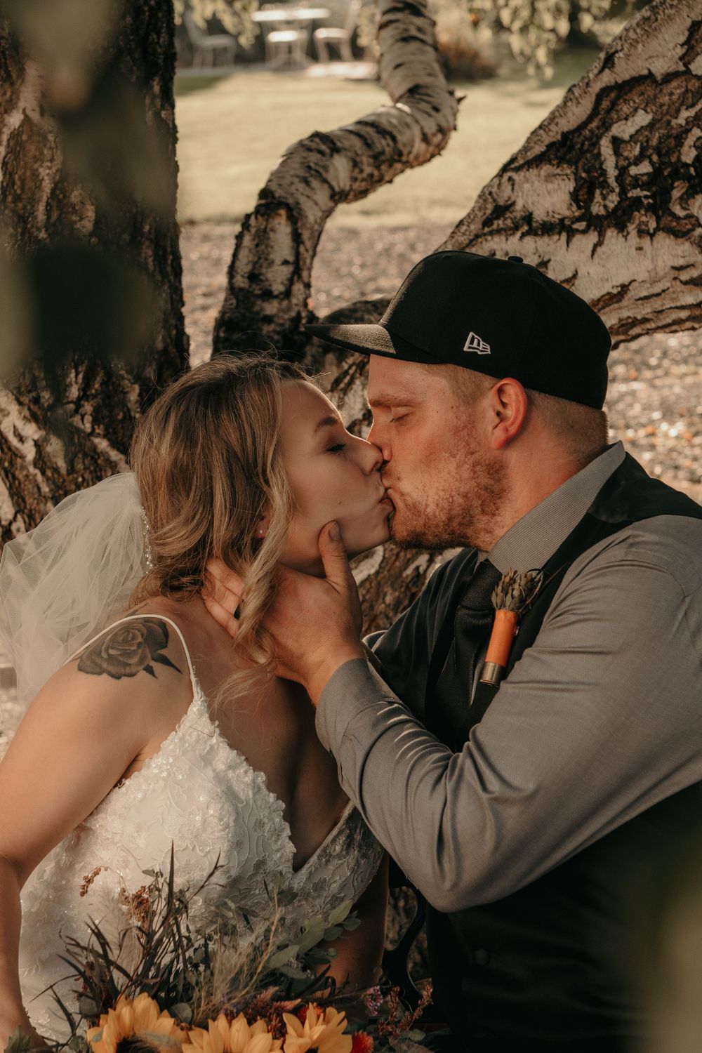 Bride and groom kissing under birch trees with sunflower bouquet and groom wearing snapback cap.