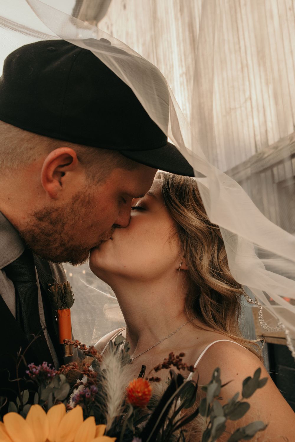 Bride and groom sharing a romantic kiss under a wedding veil with colorful bouquet in foreground.