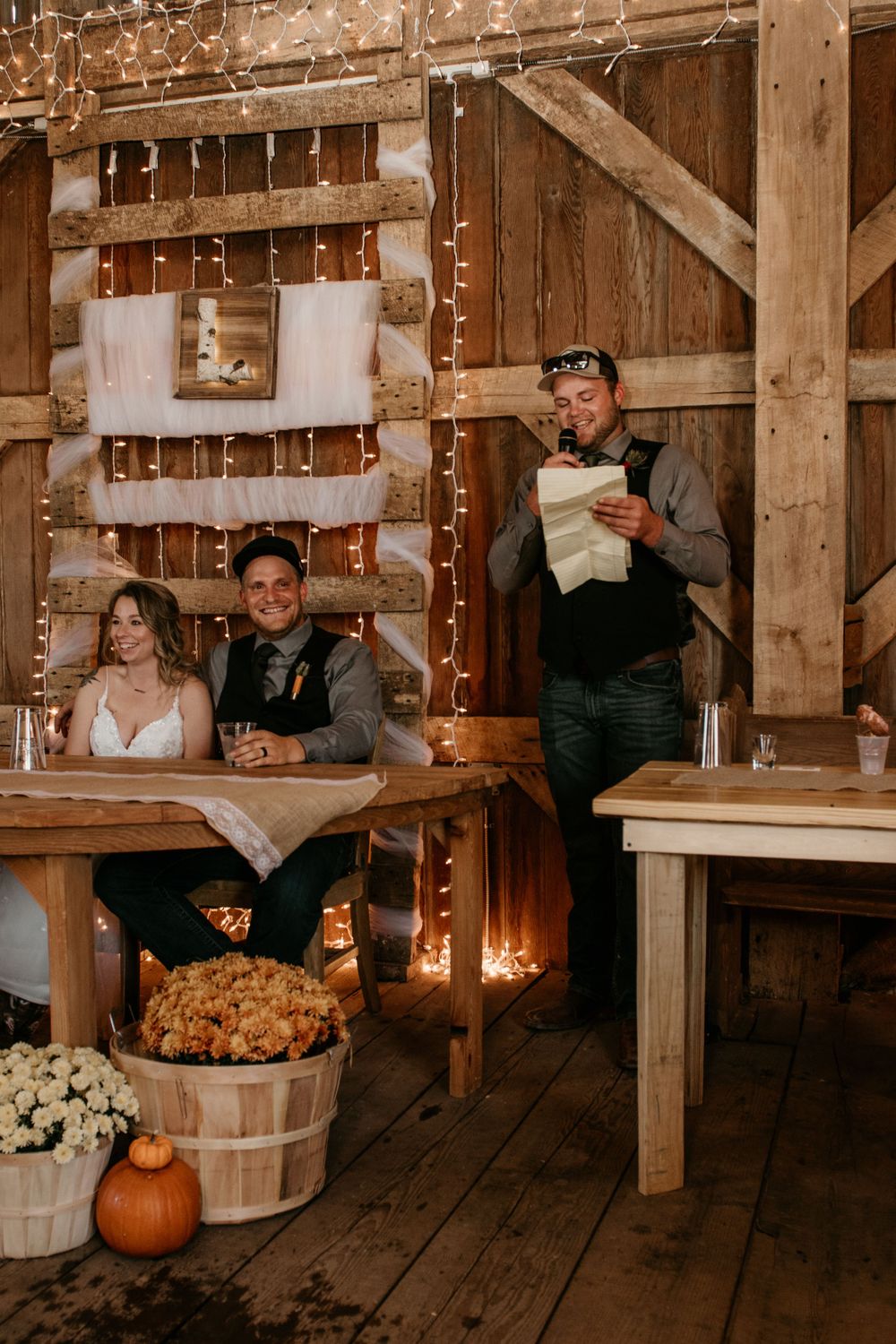 Groom reads heartfelt speech at rustic barn reception with fairy lights and harvest decor.