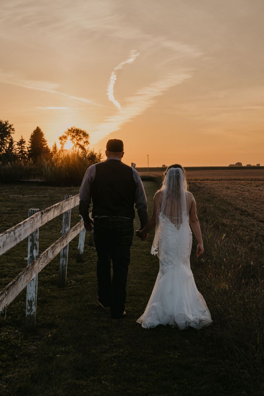 Bride and groom walk hand in hand along fence at golden sunset on rustic farm wedding.