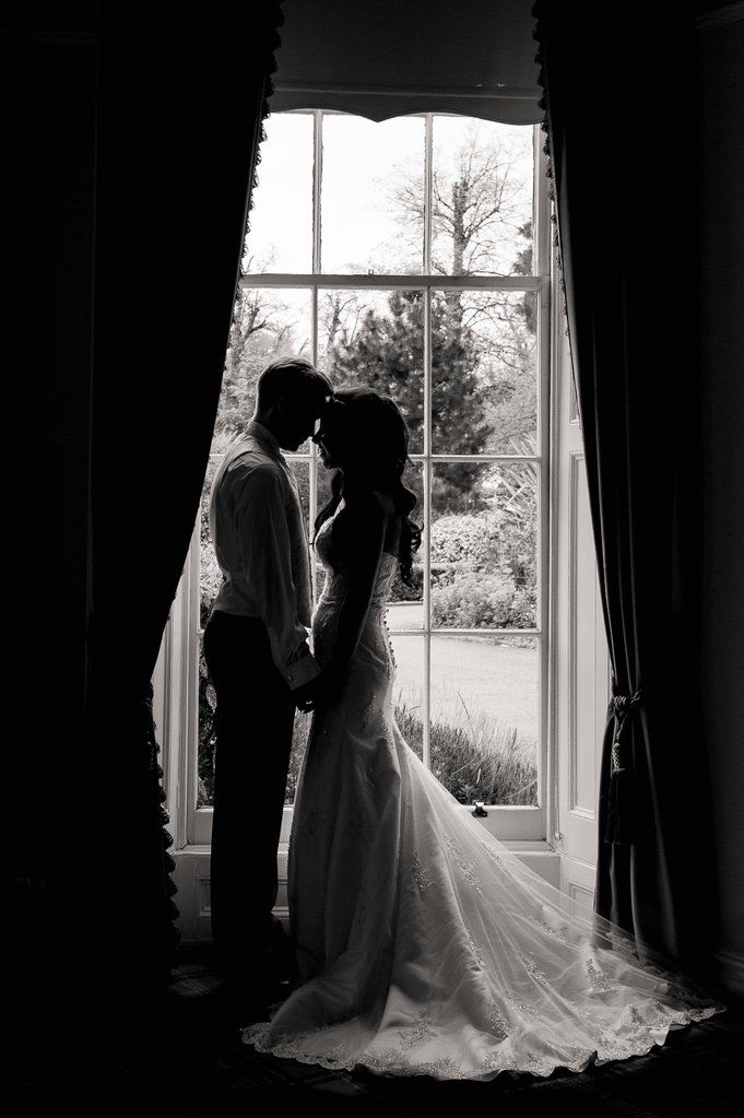 Silhouetted wedding couple embracing by a large window in black and white photography.