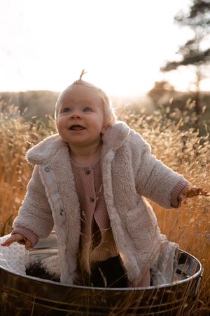 Baby in cozy sherpa coat sits in metal basin during golden hour outdoors.
