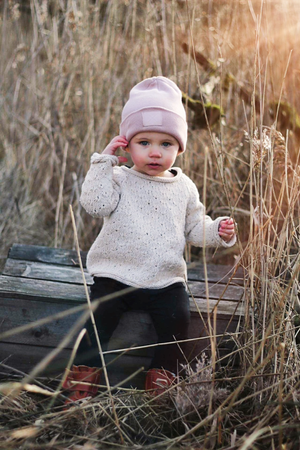 Small child in cream sweater and pink hat exploring tall grass outdoors.