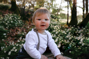 Young child in white top and suspenders sits outdoors surrounded by white wildflowers.