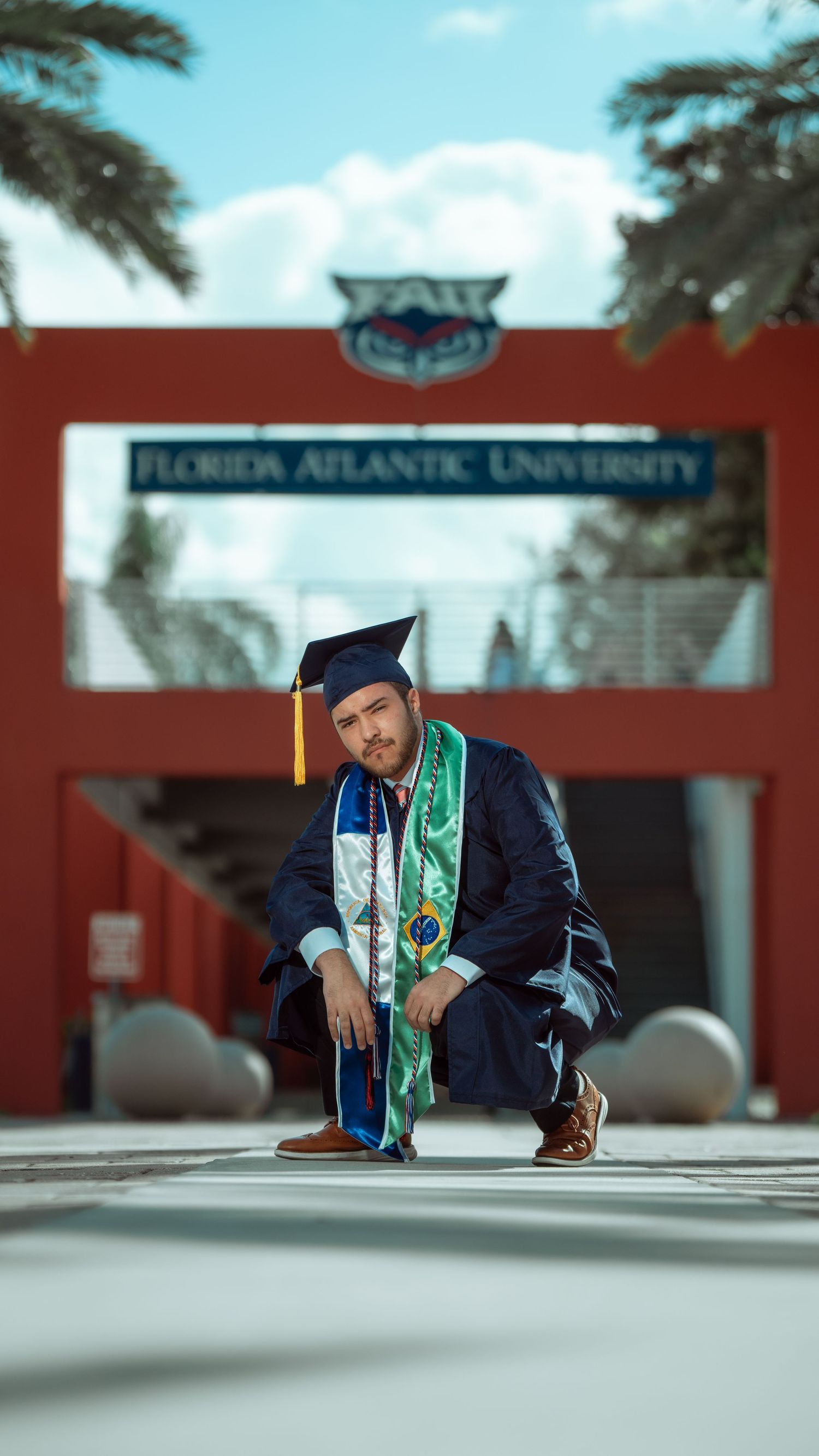 Graduate in blue cap and gown poses in front of Florida Atlantic University entrance sign.
