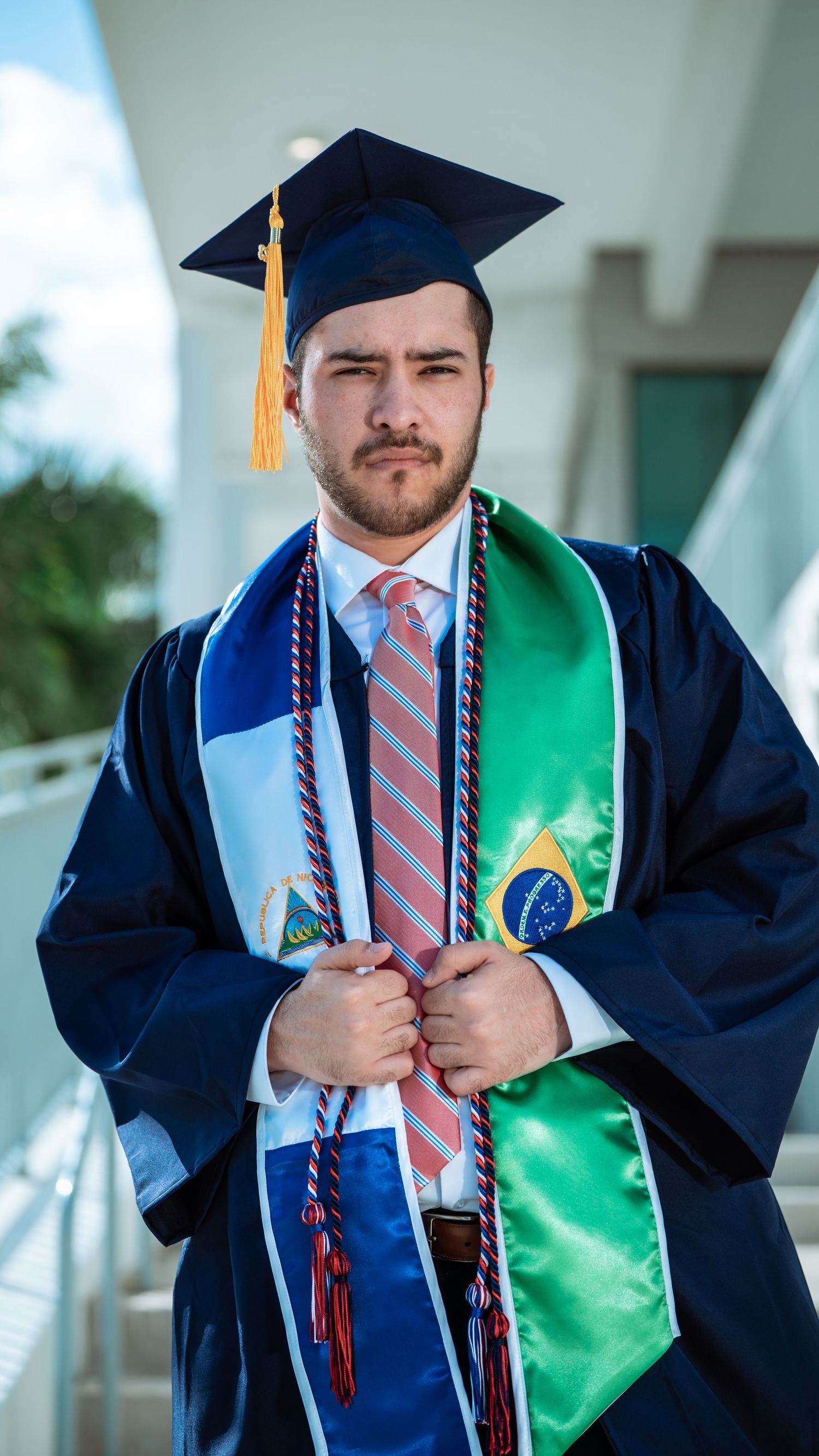 Graduate in navy cap and gown with green and blue stoles poses for portrait during commencement ceremony.