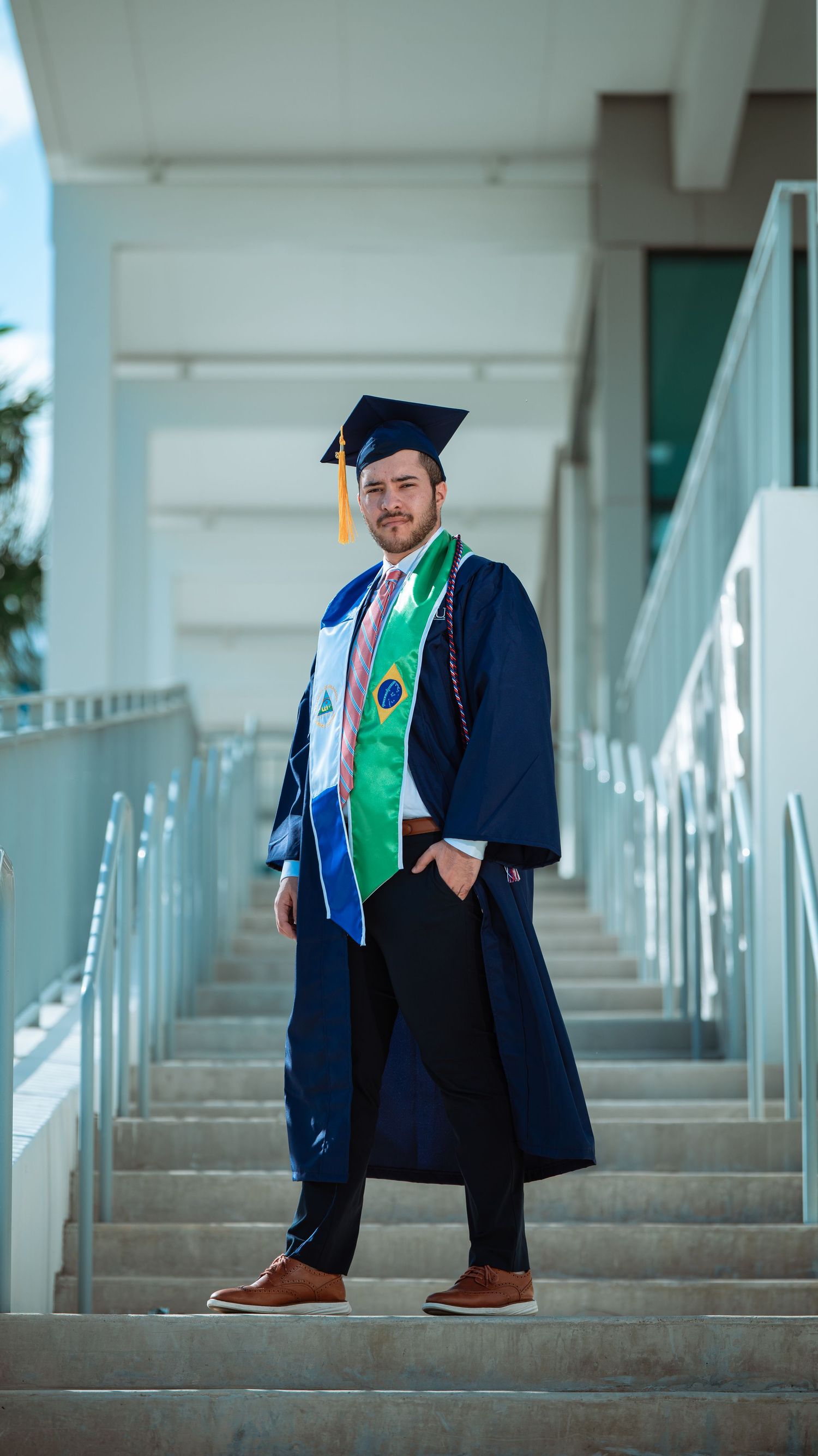 Graduate in ceremonial attire poses confidently on concrete steps wearing blue and green stoles.