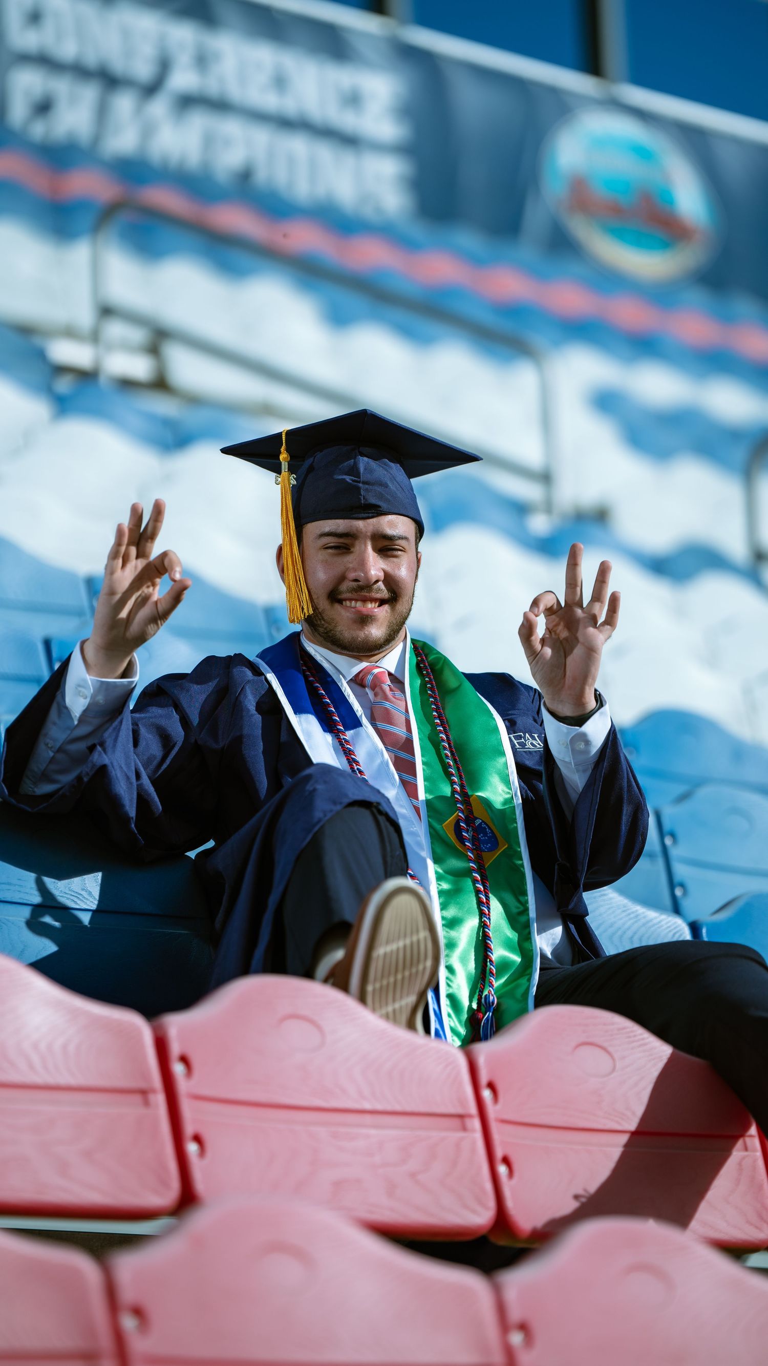 Smiling graduate raises both hands making peace signs while seated in stadium.