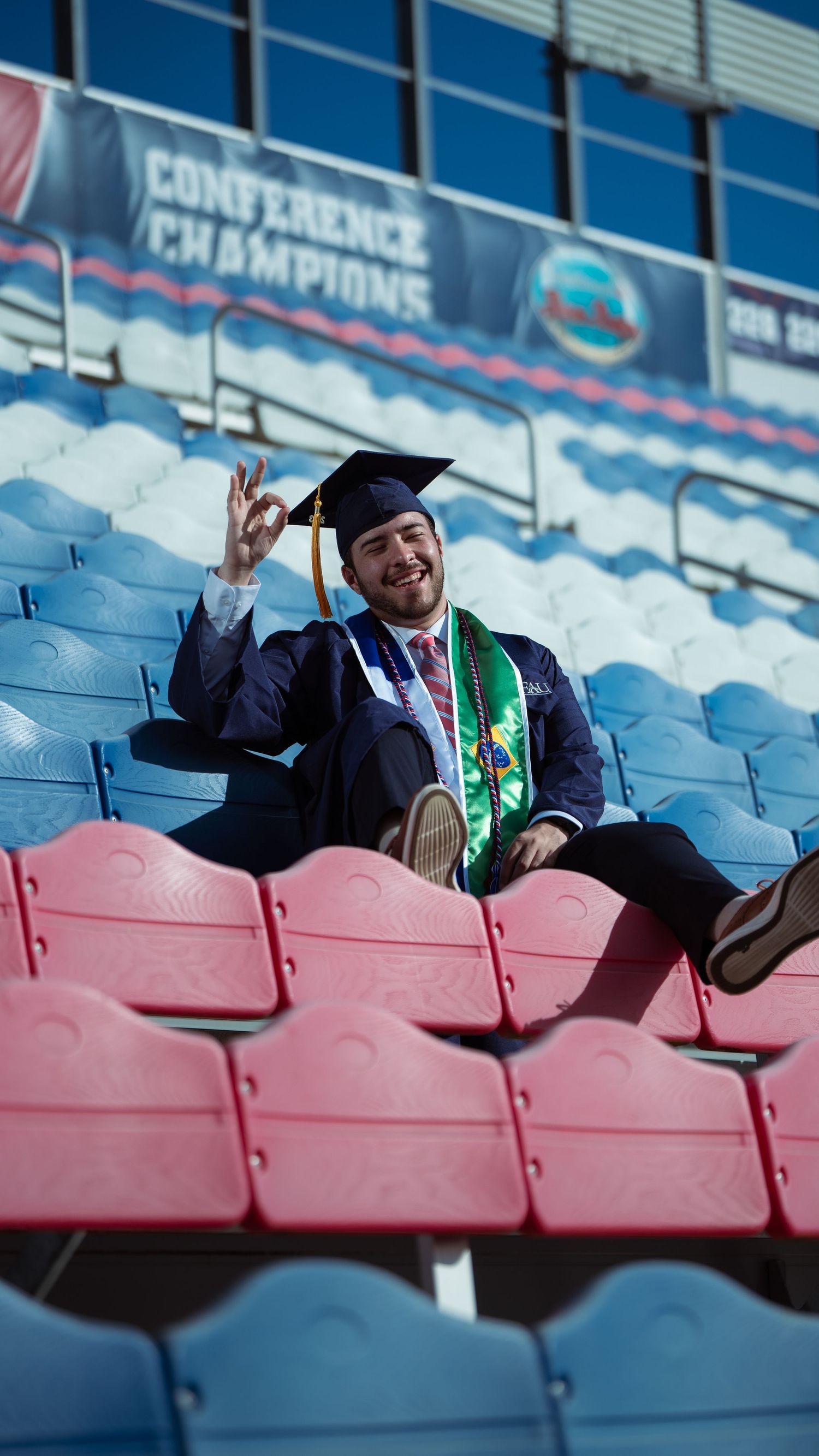 Graduate makes peace sign gesture while sitting in red stadium seats wearing cap and gown.