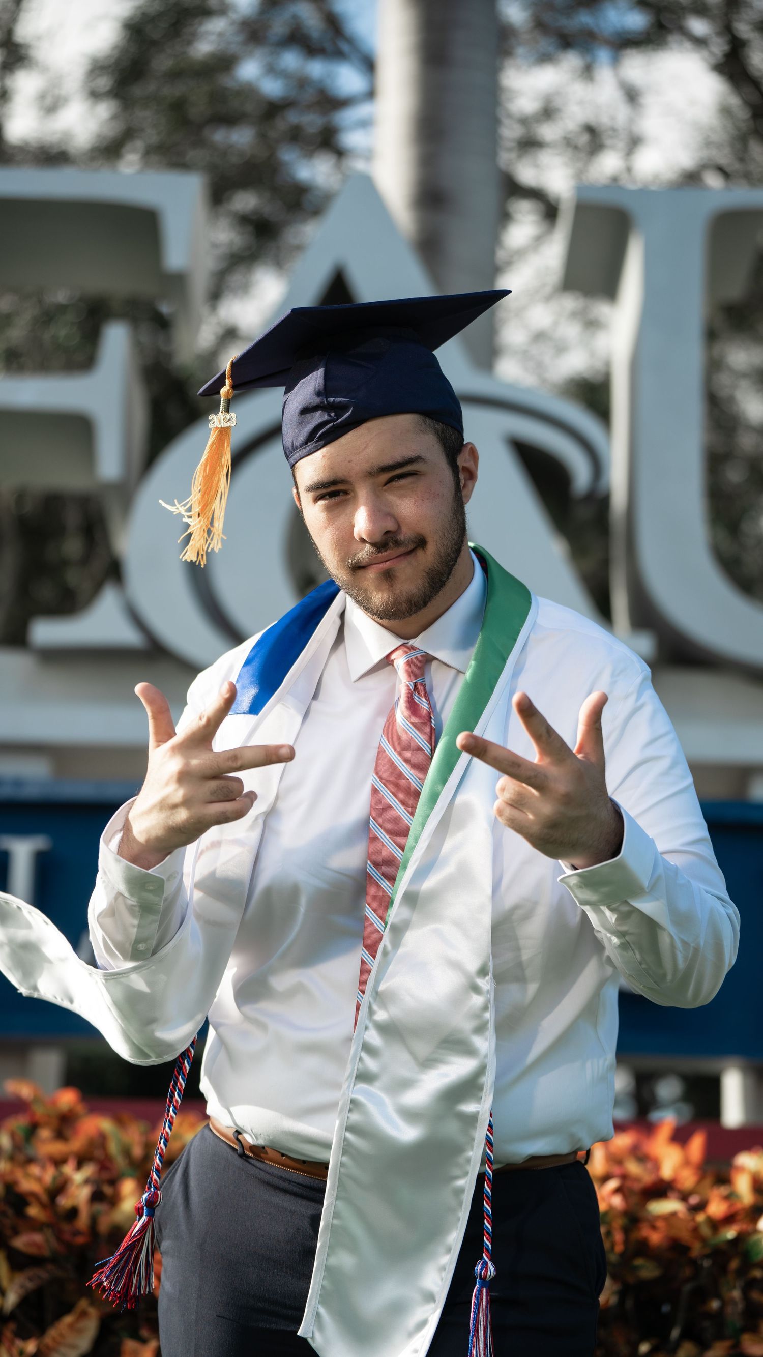 Graduate in cap and striped tie makes hand gestures during outdoor photo session.