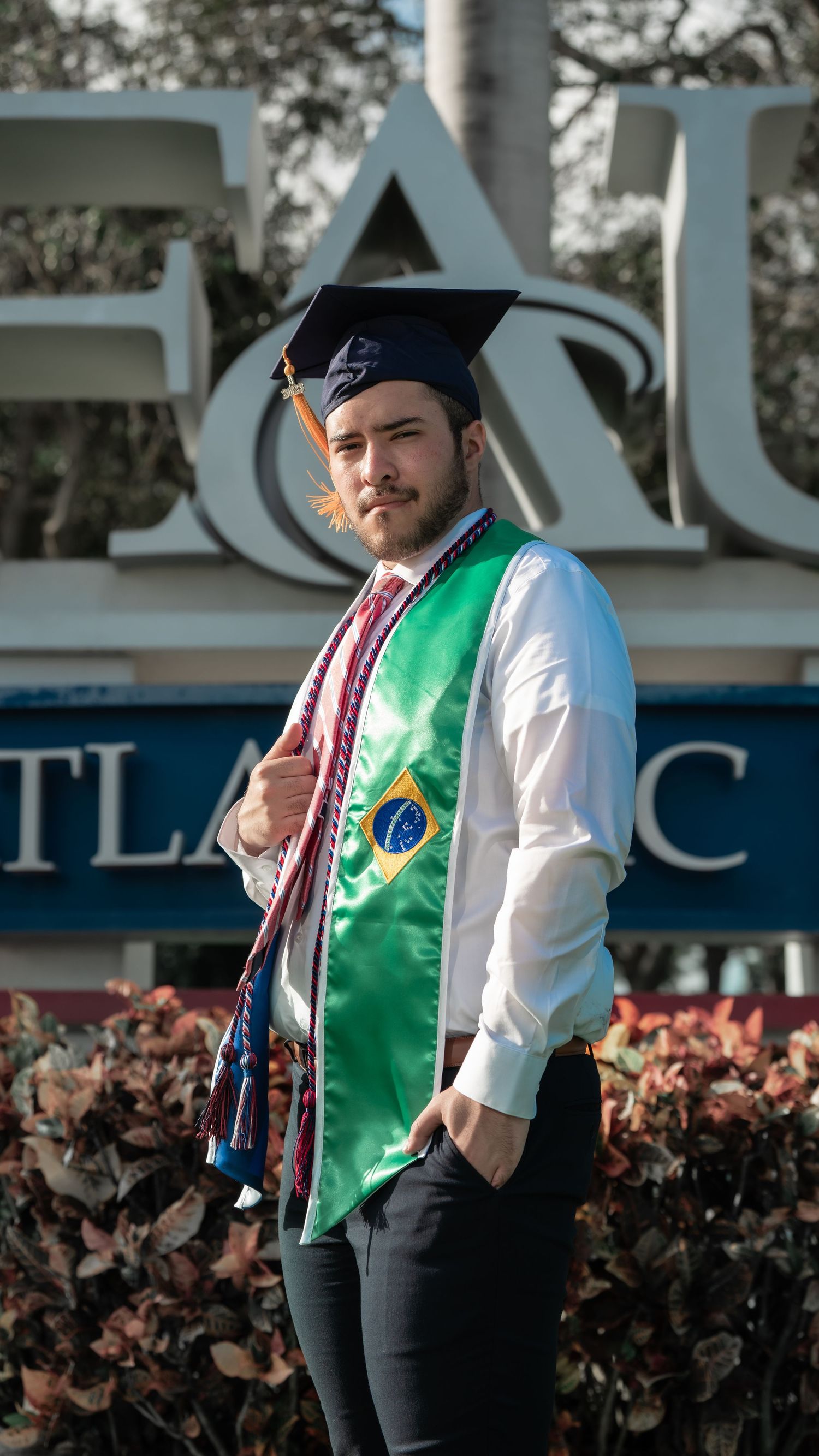 Graduate wearing Brazilian sash poses confidently in front of university sign.