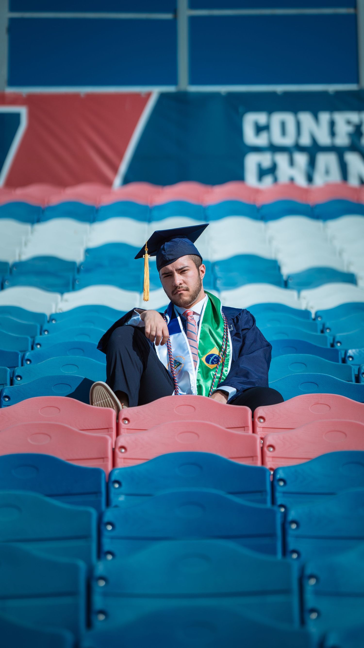 Graduate sits alone in stadium seats wearing academic regalia and green stole.