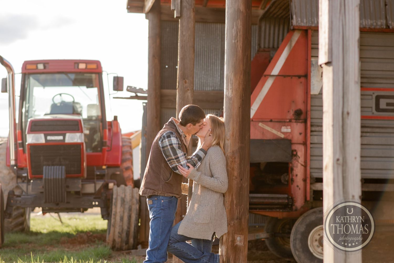 A couple sharing an intimate moment near farm equipment and a barn.