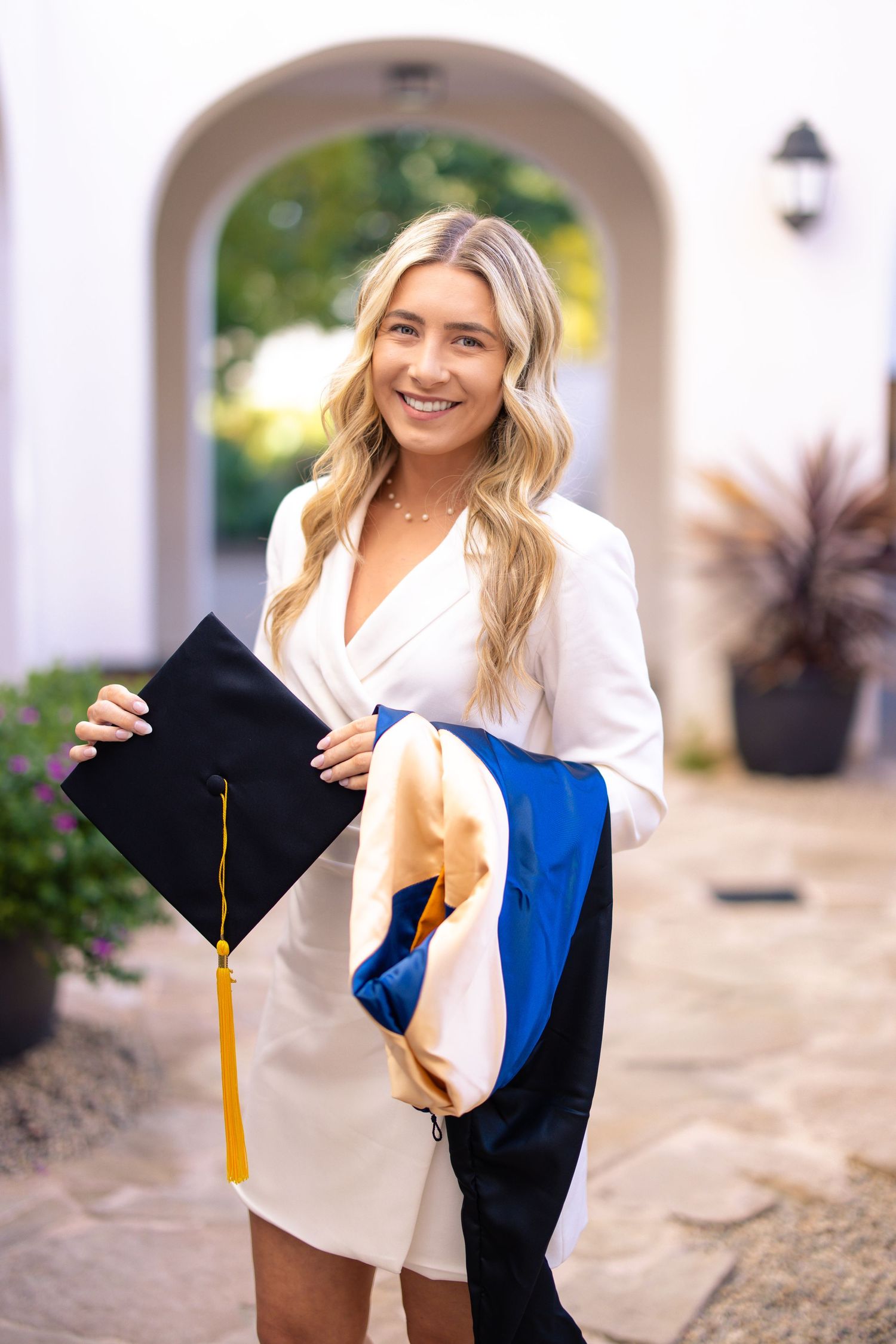 Graduate in white dress holds graduation cap with blue and gold tassel.